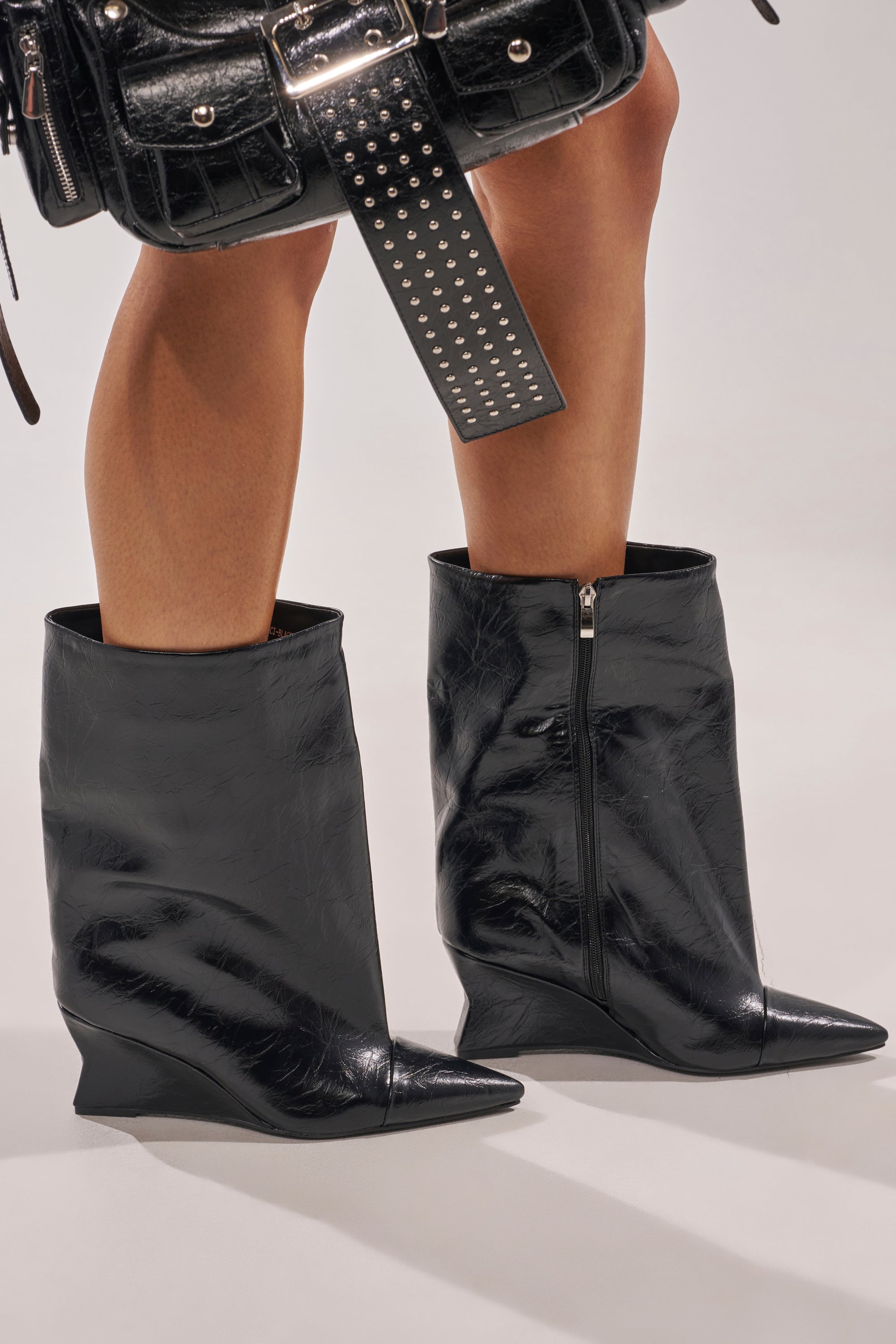 A close-up of a person wearing the AZALEA WANG VINCI BLACK PU PRINT WEDGE BOOTIE with pointed toes and side zippers, paired with a black textured handbag detailed with silver studs and hardware. 0