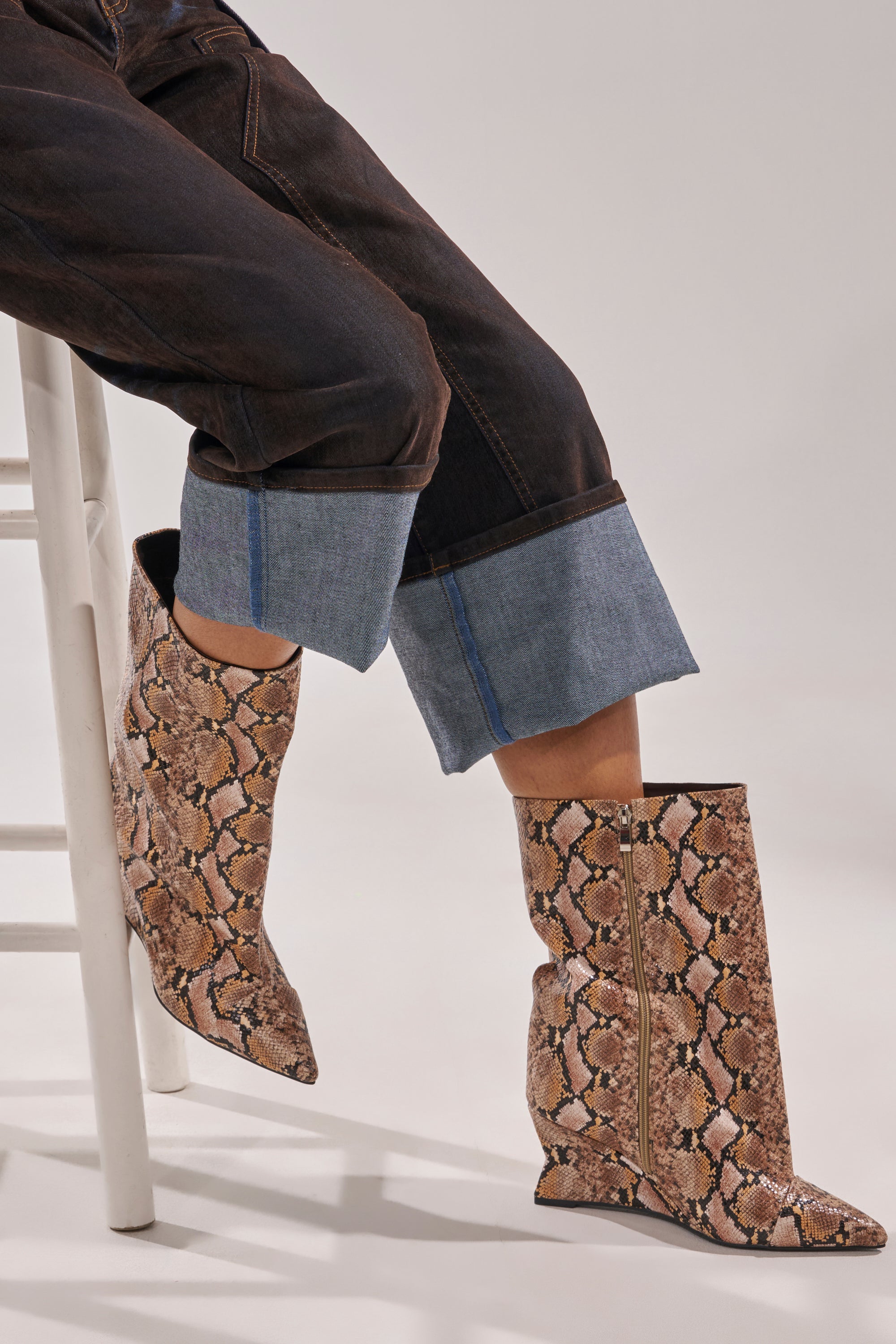 A person sits on a white stool wearing dark, cuffed jeans and the AZALEA WANG VINCI SNAKE PU PRINT WEDGE BOOTIE with a beige and brown snakeskin pattern. The background is plain and light-colored. 1