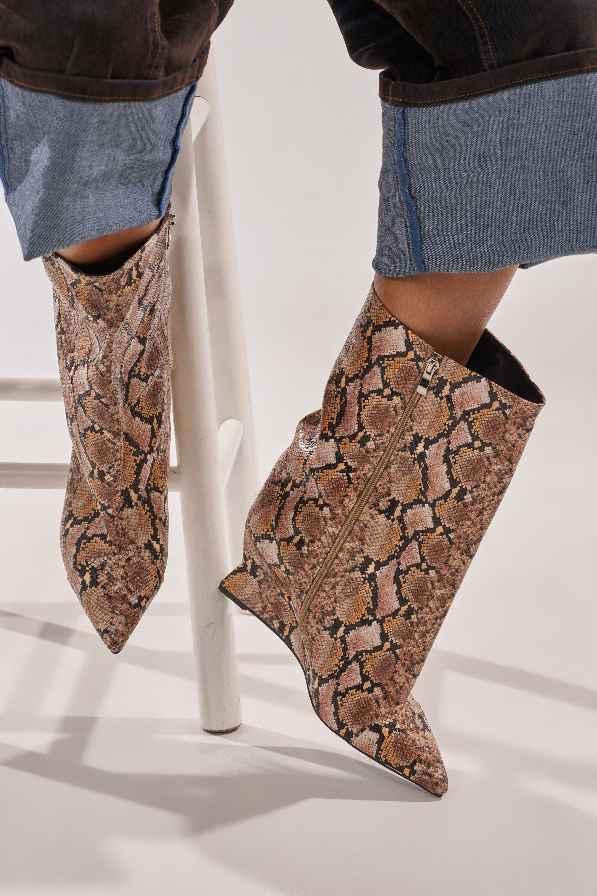 A person in blue cuffed jeans and AZALEA WANG VINCI SNAKE PU PRINT WEDGE BOOTIE sits on a white stool with legs crossed. 2