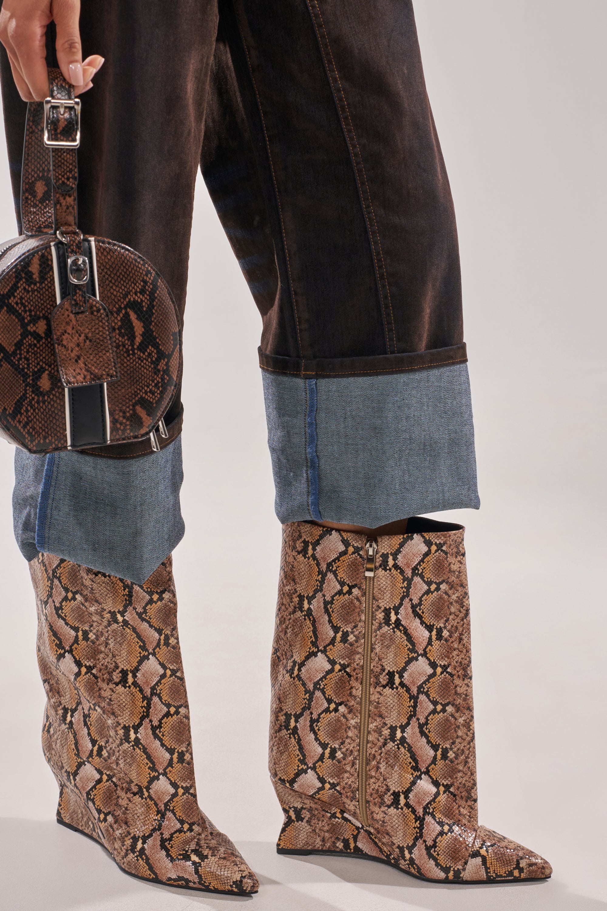 A person wears dark cuffed jeans and carries a round snakeskin handbag, styled with the AZALEA WANG VINCI SNAKE PU PRINT WEDGE BOOTIE. The background is plain and neutral. 4