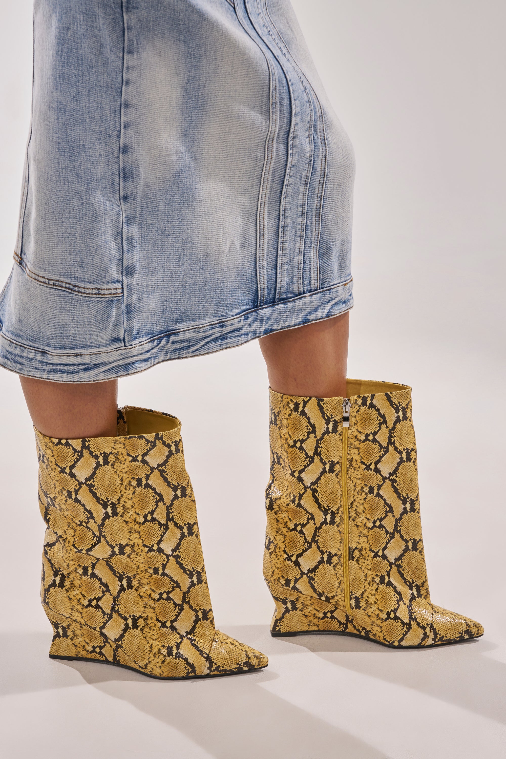 A person in a light blue denim skirt stands on a light background wearing AZALEA WANG VINCI Yellow Snake PU Print Wedge Bootie. 0