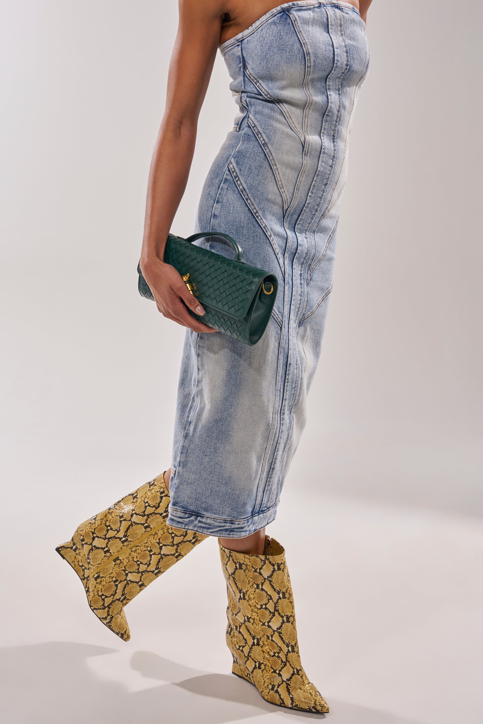 A person in a strapless denim dress and textured green clutch stands against a plain light background, wearing the AZALEA WANG VINCI YELLOW SNAKE PU PRINT WEDGE BOOTIE. 1