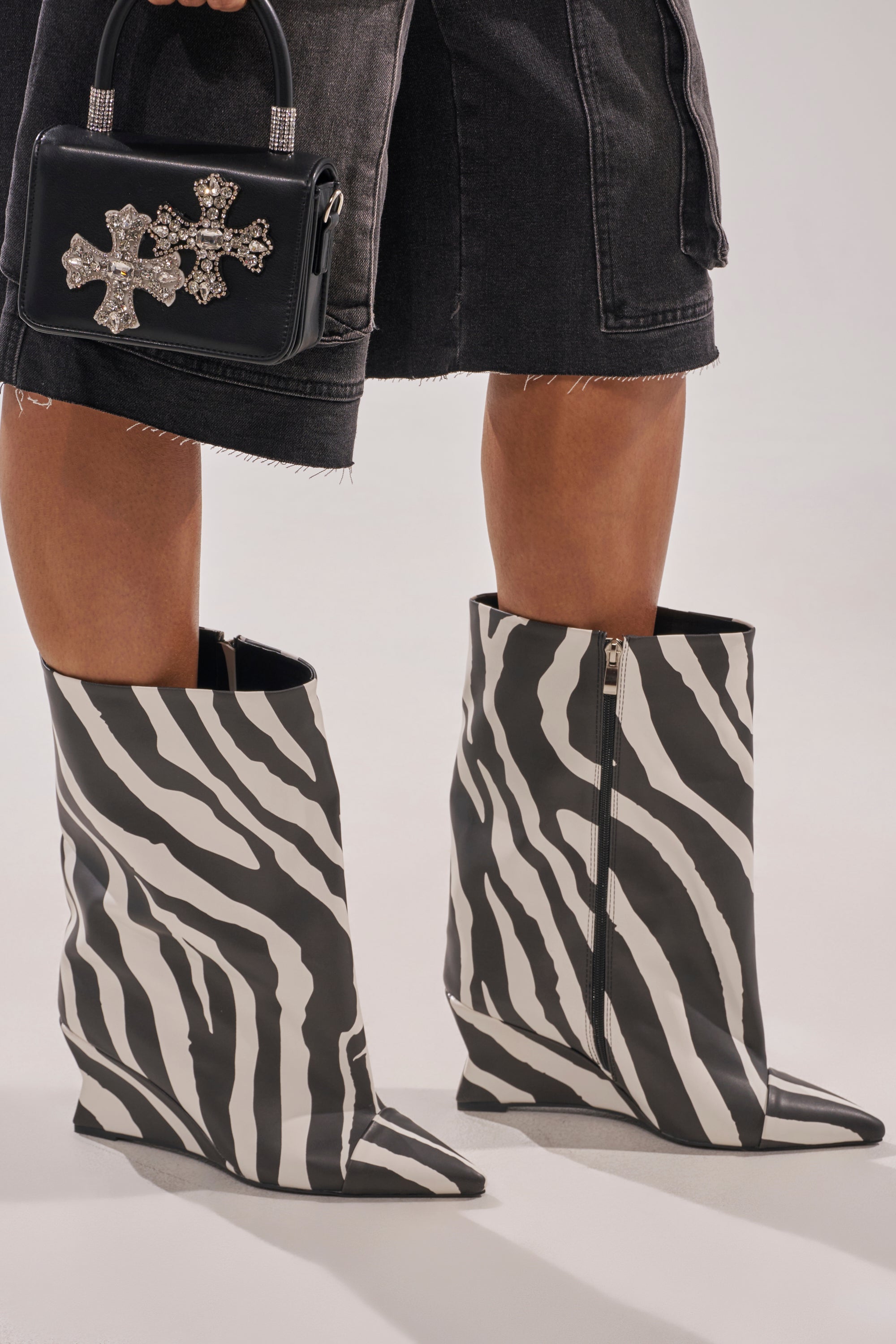 Wearing the AZALEA WANG VINCI ZEBRA PU PRINT WEDGE BOOTIE and a black denim skirt, a person holds a small black handbag adorned with jeweled cross embellishments. 0