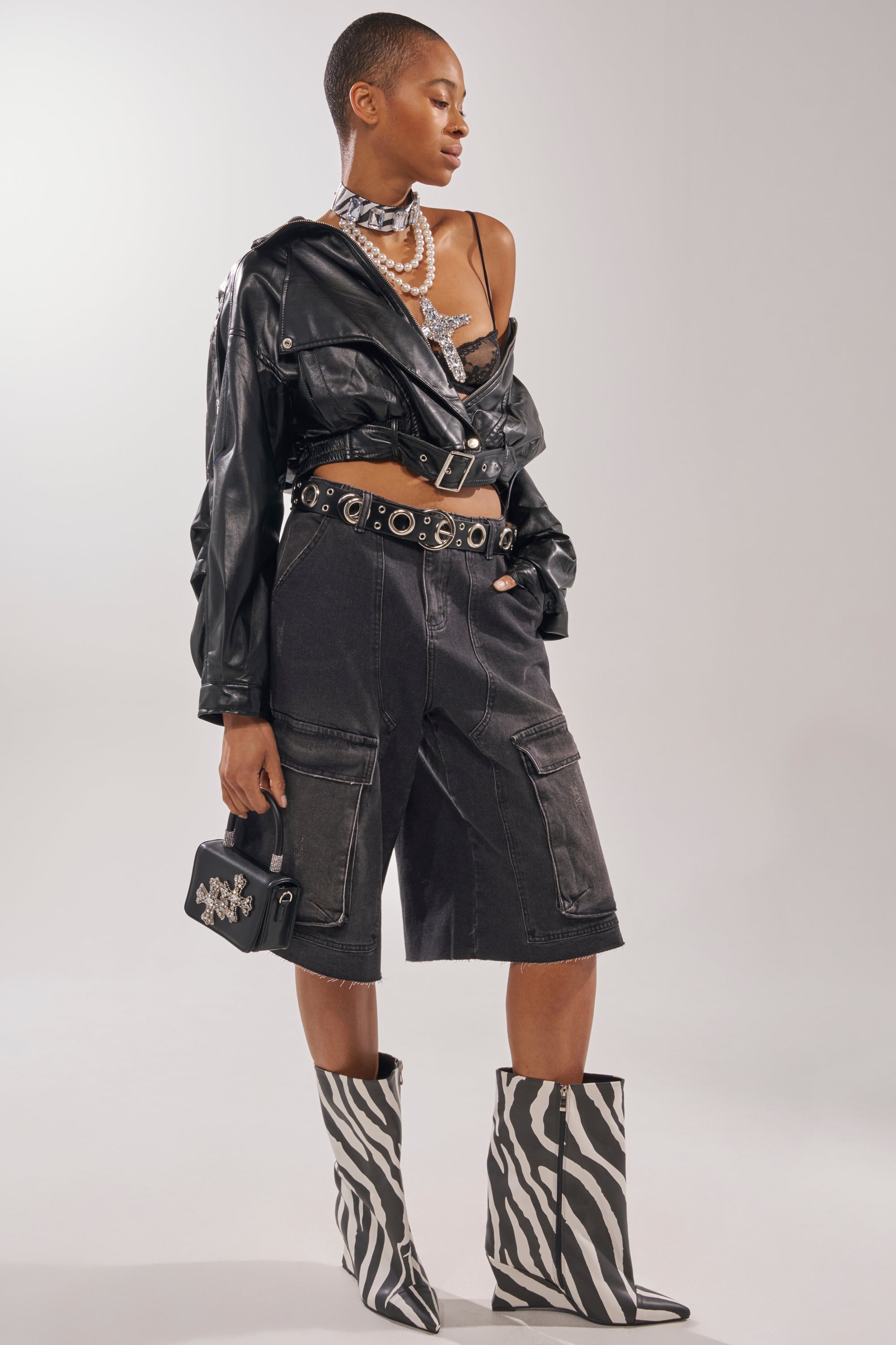 A person wearing a black leather jacket over a lace bralette, knee-length black cargo shorts, layered necklaces, a studded belt, holding a black handbag with silver details, and AZALEA WANG VINCI ZEBRA PU PRINT WEDGE BOOTIE. 1