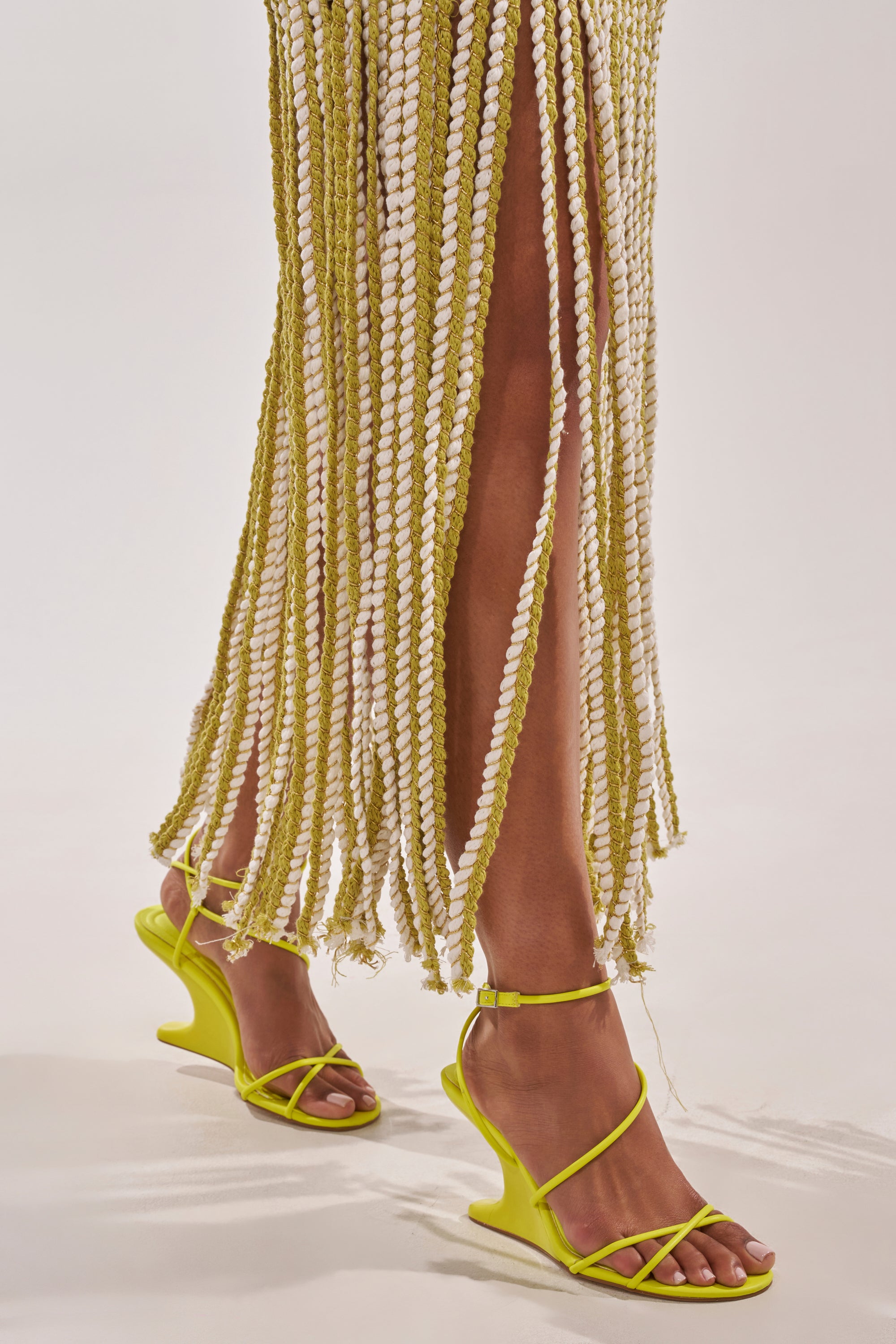 A person wears a skirt of twisted white and yellow-green rope strands, paired with the AZALEA WANG VINTARA YELLOW NOVELTY WEDGE STRAP SANDAL, standing on a plain white background. 1