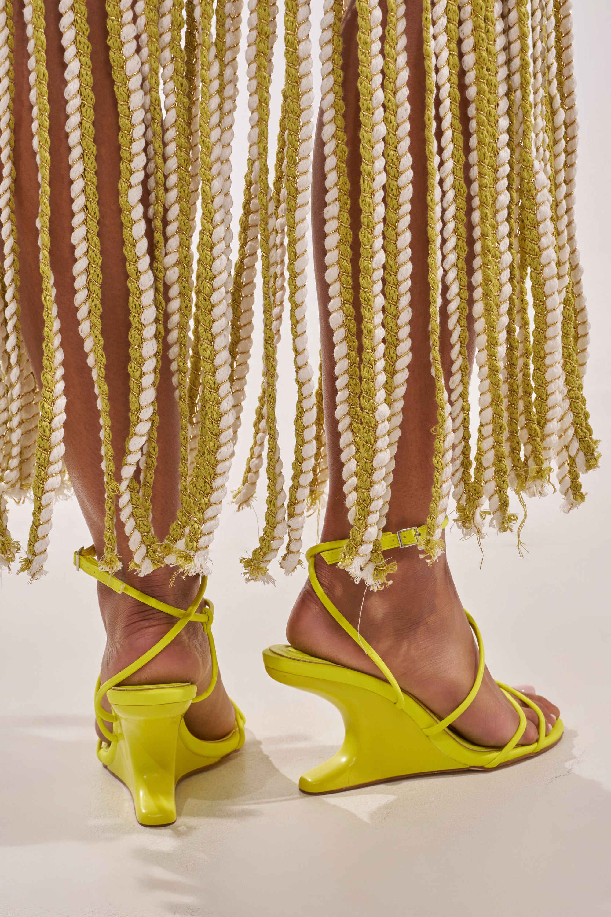 A person stands on a light surface wearing AZALEA WANG VINTARA YELLOW NOVELTY WEDGE STRAP SANDALS, paired with a skirt or dress featuring long, thick, twisted yellow and white fringe around their legs. 3