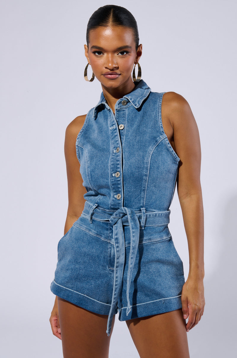 A person poses in the SAYLOR DENIM ROMPER, a sleeveless piece featuring a belted waist and button-down front. They have their hair tied back and are adorned with large hoop earrings, gazing directly at the camera against a plain light-colored background.