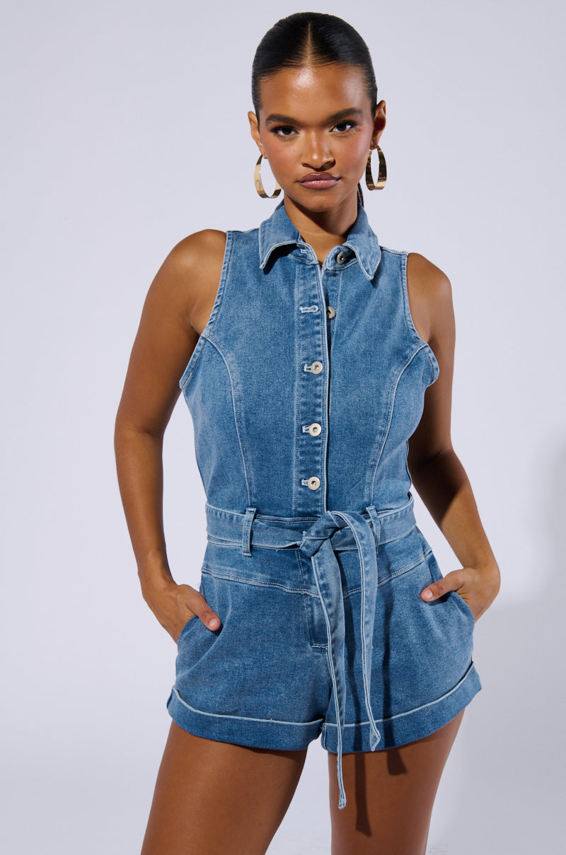 A woman stands confidently against a white background, wearing the SAYLOR DENIM ROMPER with its sleeveless design, button-down front, and tied waist belt. She has her hands in the pockets of her shorts and accessorizes with large hoop earrings.