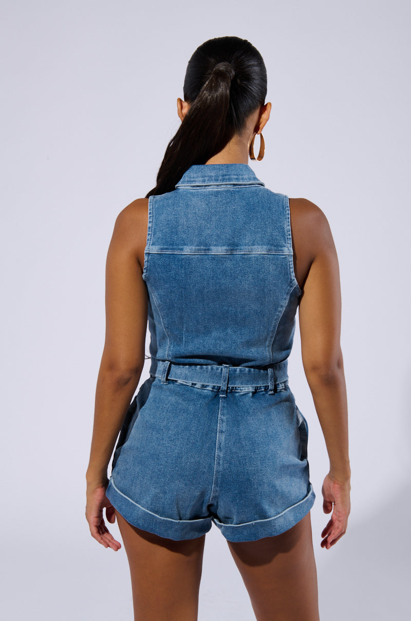 A person with long, dark hair in a ponytail is standing with their back to the camera, showcasing the SAYLOR DENIM ROMPER. The outfit features a sleeveless design with a collar, belt, and button-down front closure. They are accessorized with large hoop earrings against a plain, light-colored background.