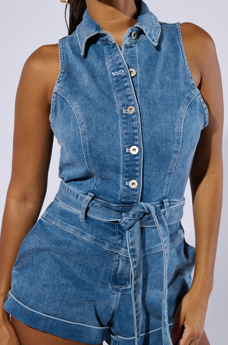 A person wearing the SAYLOR DENIM ROMPER, which features a sleeveless design, button-down front, and a tie belt at the waist. The romper includes a collar, seam detailing, and rolled hem shorts. The background is plain, and the person's head is not visible in the image.