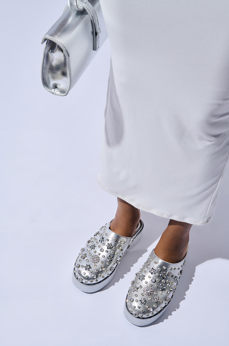 A person wearing the AZALEA WANG VIRTUOUS SILVER FLATFORM shoes, adorned with rhinestones and featuring a slip-on fit, paired with a white dress, stands on a light background. They are holding a shiny silver clutch bag. 0