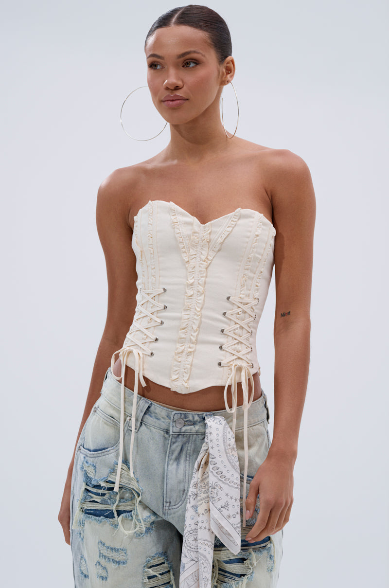 A woman wears the LETS GET ROWDY LACE UP CORSET in cream, styled with light blue distressed jeans and a patterned bandana at her waist, standing against a plain light background. 0