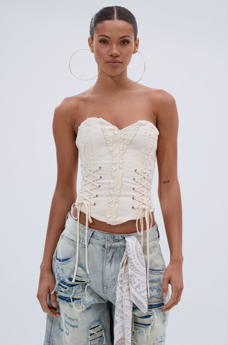 A woman wearing the LETS GET ROWDY LACE UP CORSET, large hoop earrings, distressed light-wash jeans, and a white patterned scarf tied at the waist stands against a plain background. 1