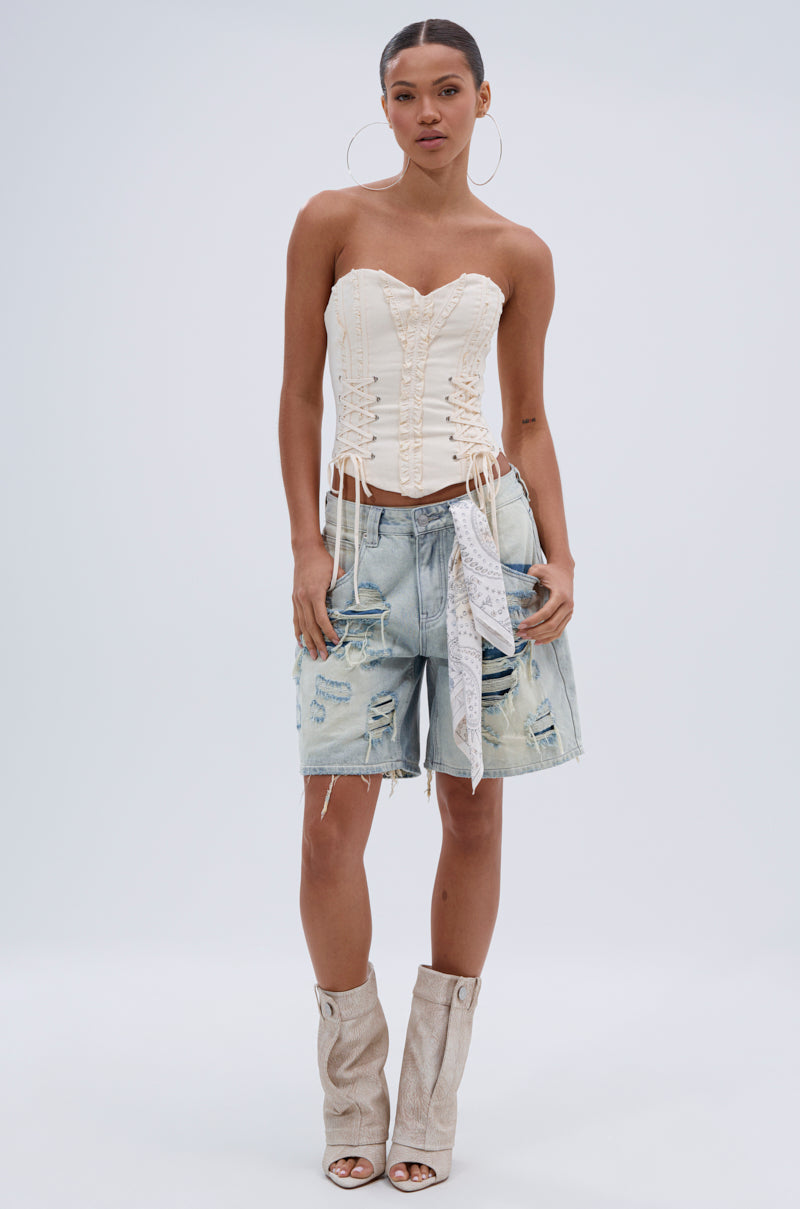 A woman stands against a plain background wearing the LETS GET ROWDY LACE UP CORSET in cream, paired with distressed denim Bermuda shorts, a white bandana in the pocket, and slouchy beige open-toe boots. 4