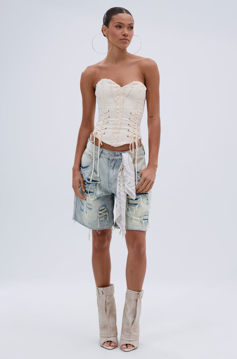 Woman wearing the LETS GET ROWDY LACE UP CORSET in cream, paired with distressed denim knee-length shorts, a white bandana, and beige open-toe heeled boots, standing against a plain light background. 5