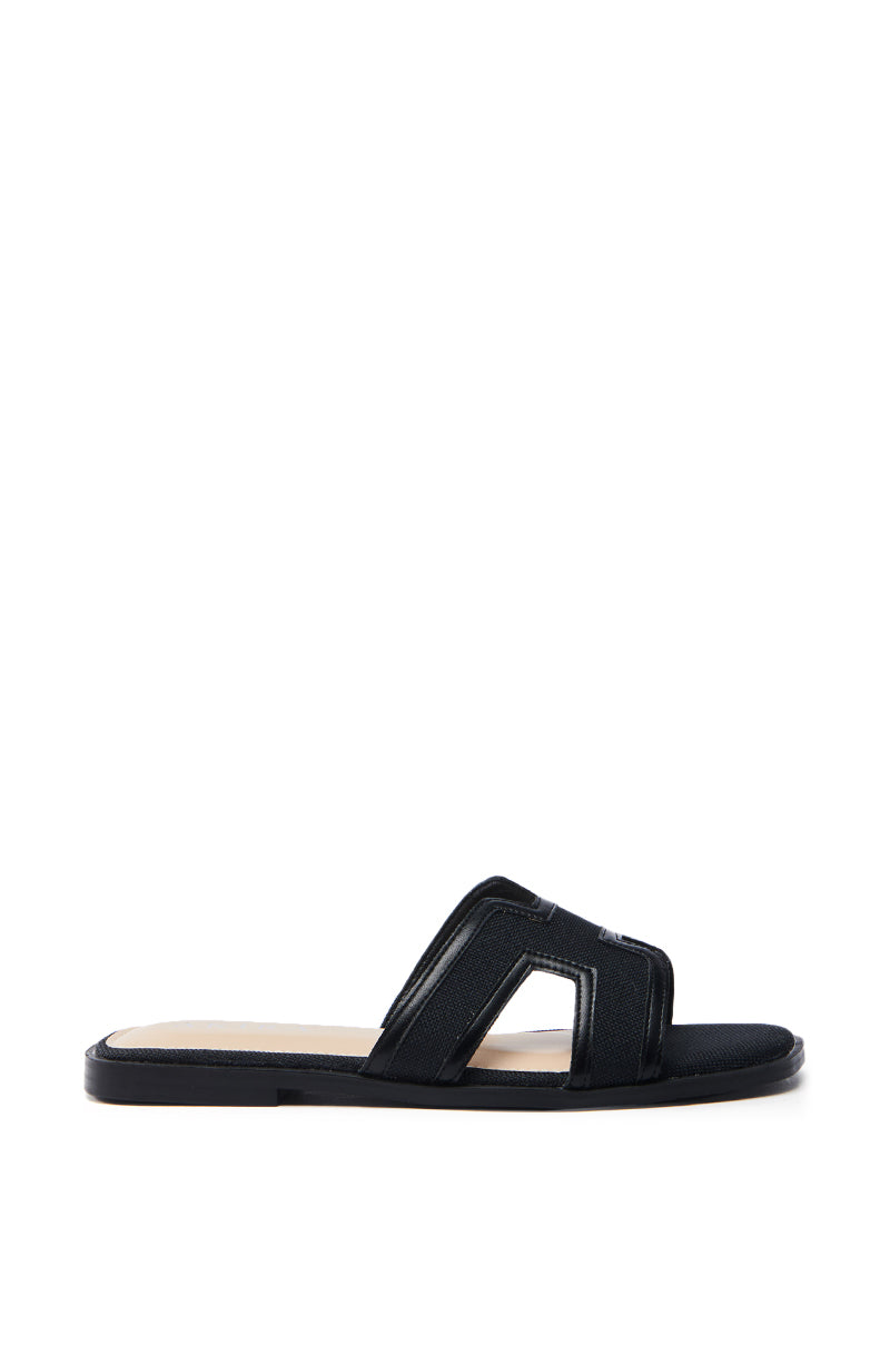 The AKIRA Vittoria Black Sandal is a black faux leather slide with a flat sole and a unique cutout on the wide strap. Its light tan insole contrasts with the black exterior, creating a minimalist and elegant look suitable for any occasion.