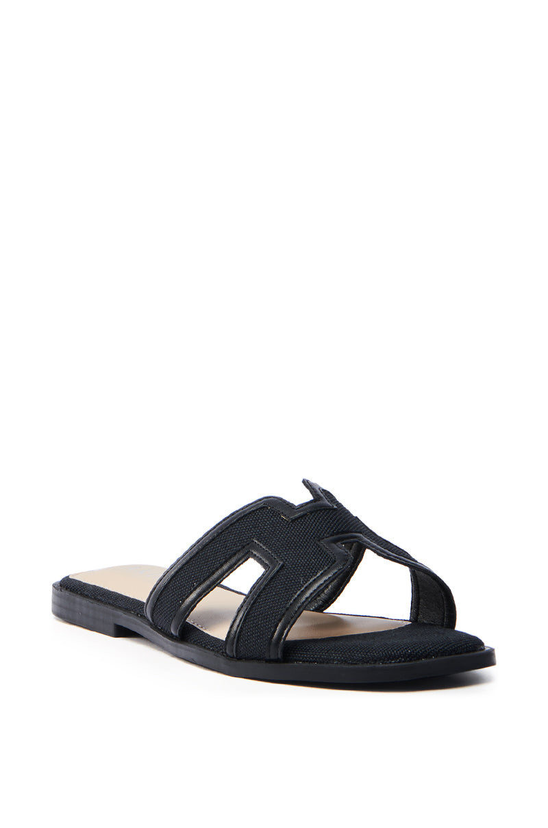 The AKIRA VITTORIA BLACK SANDAL is a sleek black slide with a flat sole and faux leather cut-out straps featuring a bold zigzag pattern, showcased with an angled view highlighting both the side and front.