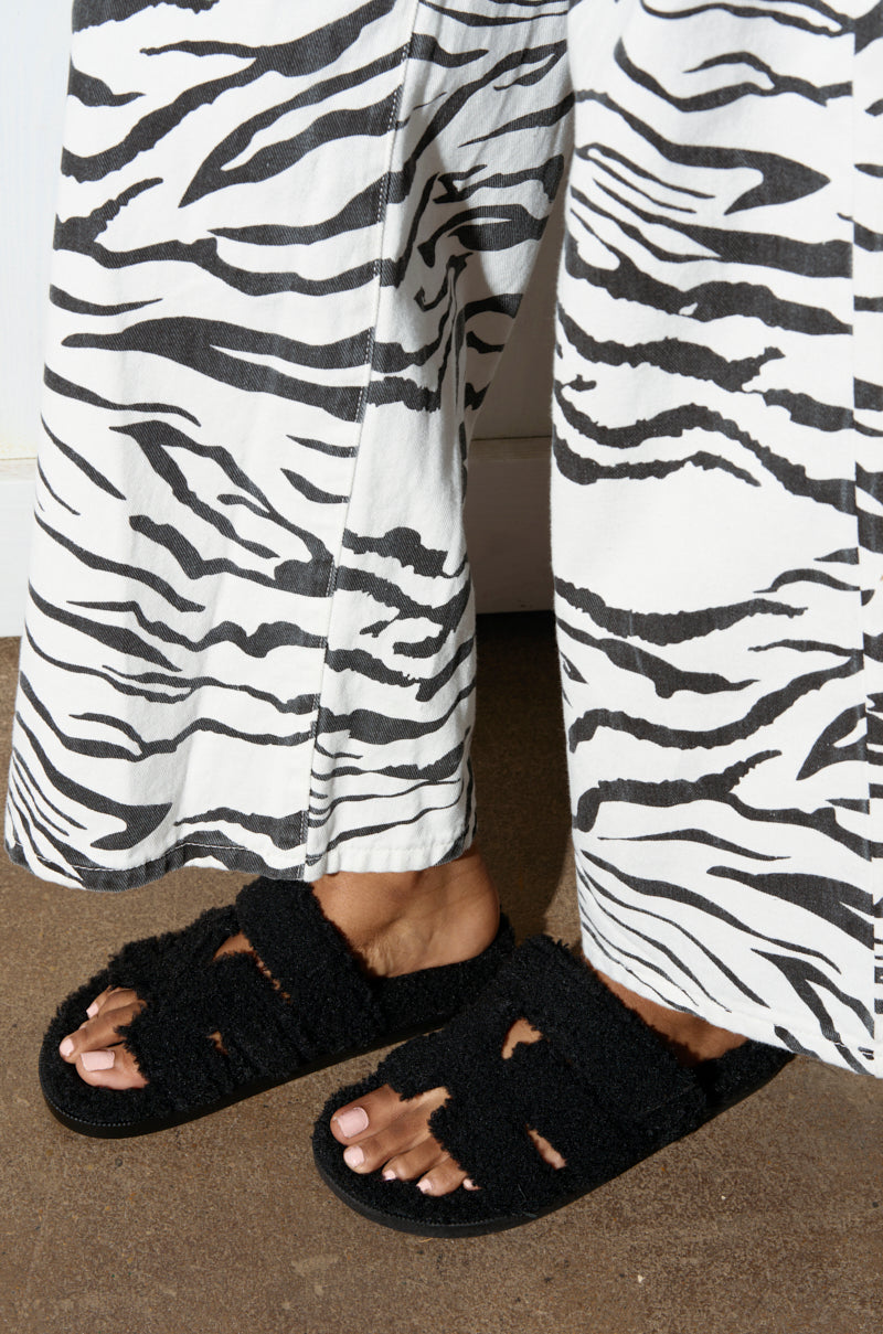 Feet adorned in the AZALEA WANG VIVEK BLACK FUZZY SLIP ON SANDAL, made from sherpa-inspired textiles, are placed on a concrete floor. The individual is wearing pants with a bold black and white zebra print pattern.