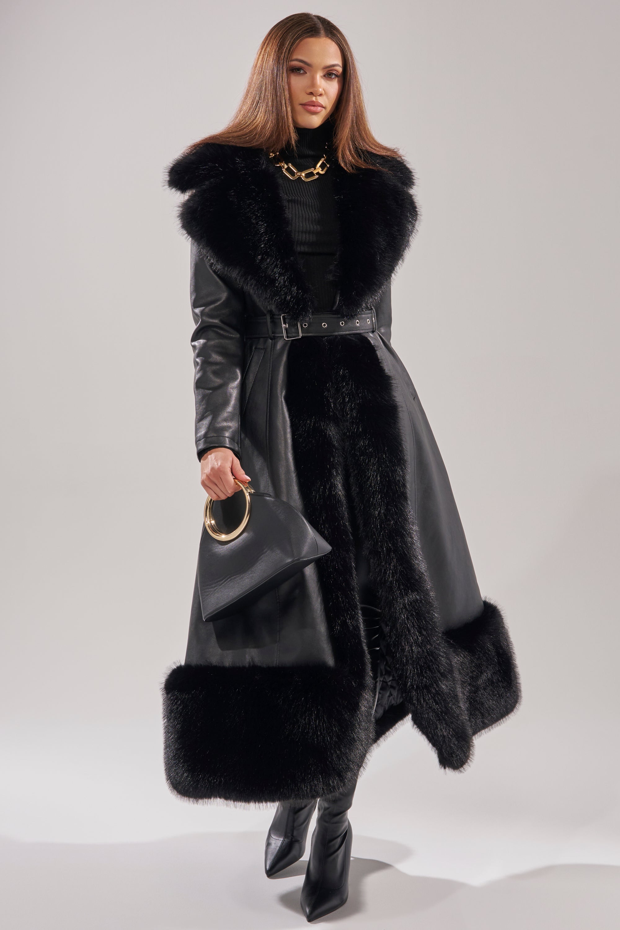 A woman exudes confidence in the VIVAN LONG PU TRENCH WITH FAUX FUR TRIM, paired with black boots, a black handbag with a gold handle, straight brown hair, and a bold chunky gold necklace.