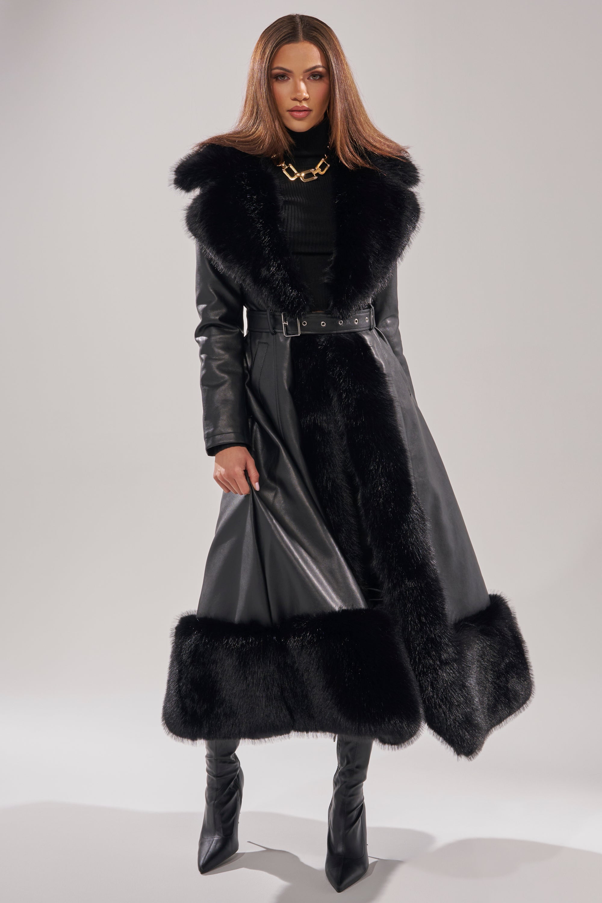A woman models the VIVAN LONG PU TRENCH WITH FAUX FUR TRIM—featuring black fur accents on the collar and hem—paired with black boots, a turtleneck, and a chunky gold necklace, against a plain light background.