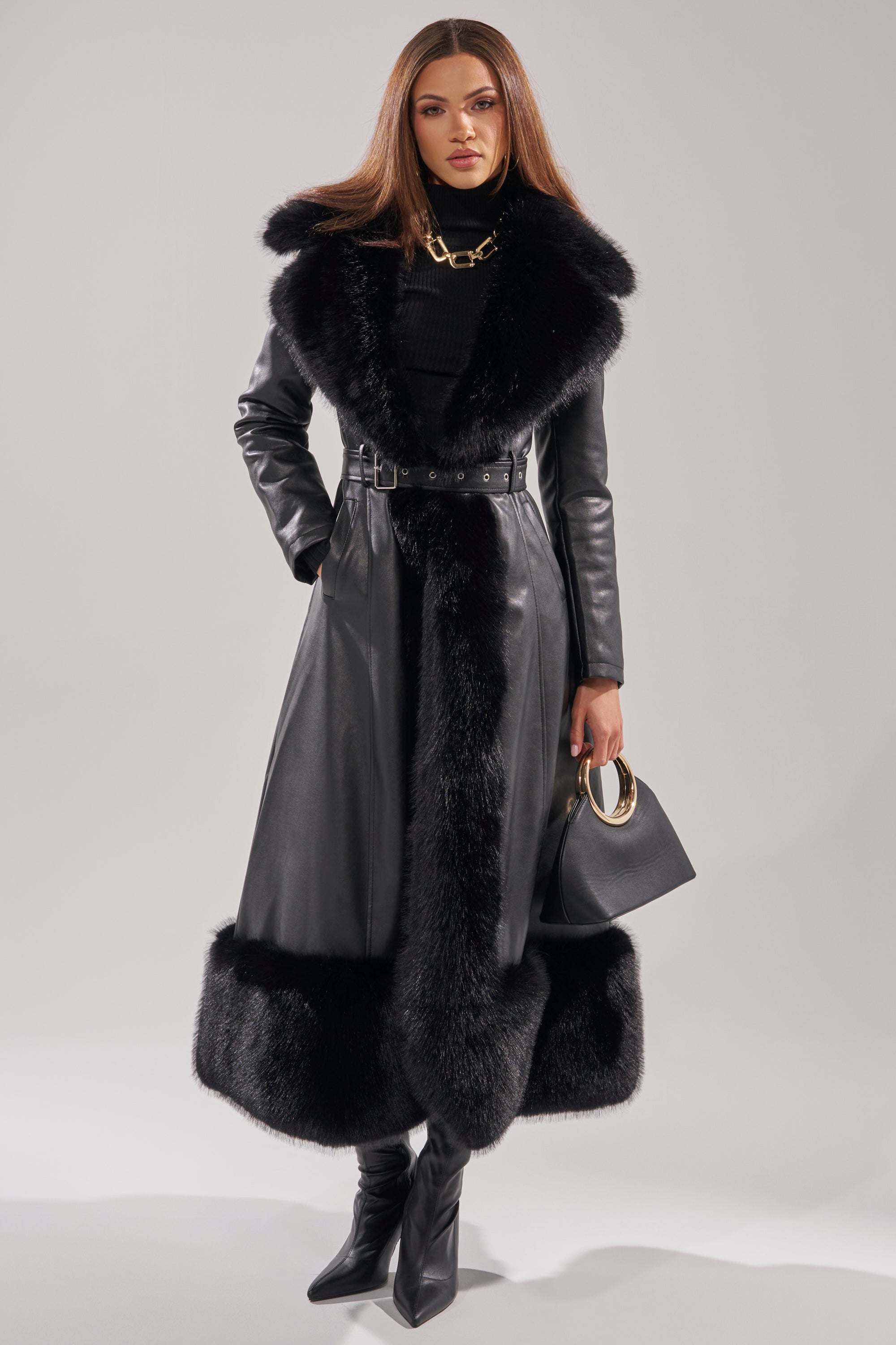 A woman wears the VIVAN LONG PU TRENCH WITH FAUX FUR TRIM, styled with a black turtleneck, heeled boots, a chunky gold necklace, and carries a black handbag with a gold handle.