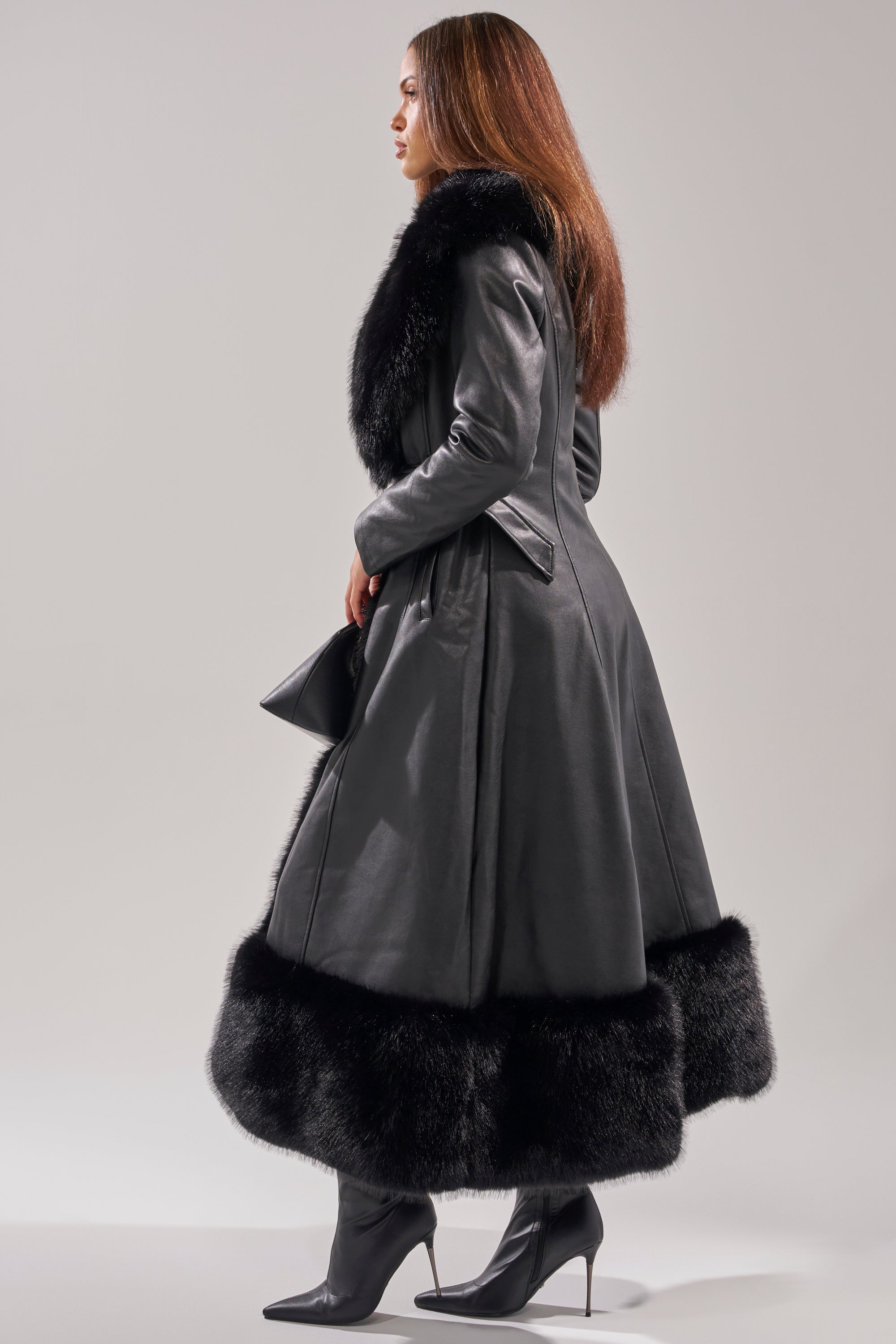 A woman stands sideways against a plain background wearing the VIVAN LONG PU TRENCH WITH FAUX FUR TRIM, paired with black heeled boots and holding a small black clutch.