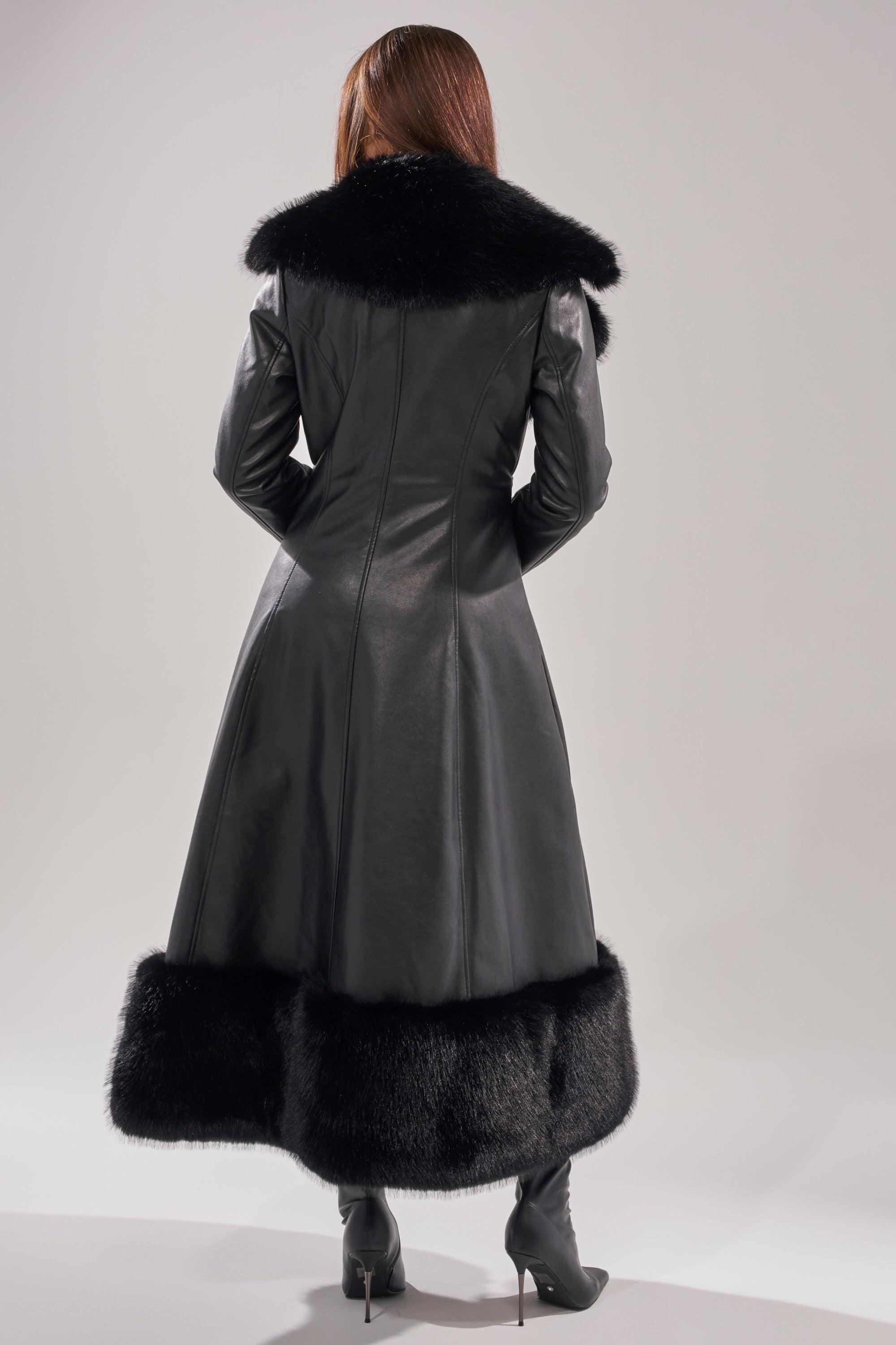 A person with long brown hair, seen from the back, wears the VIVAN LONG PU TRENCH WITH FAUX FUR TRIM and black heeled boots, standing against a plain light background.
