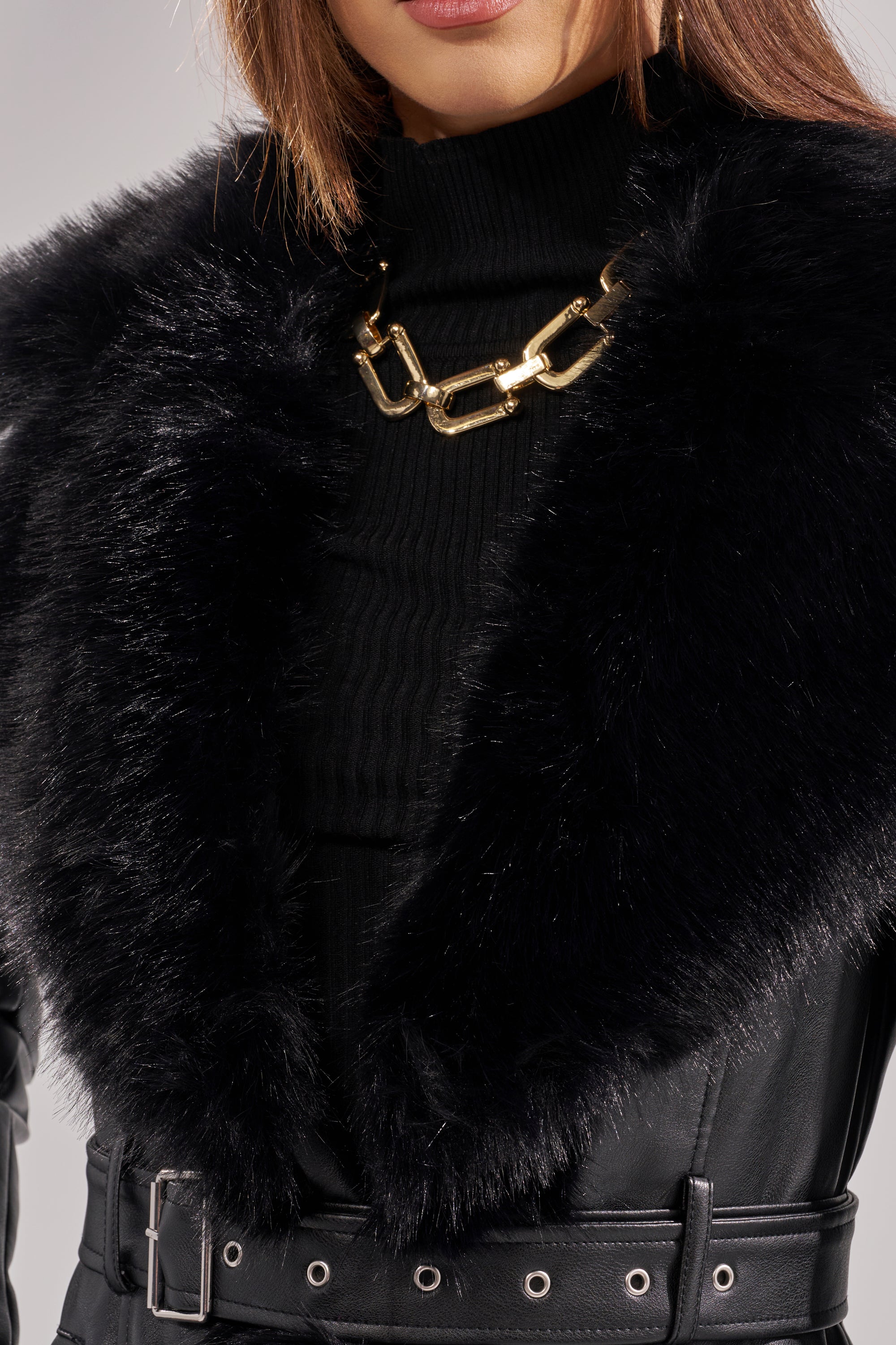 A person wears a textured black turtleneck, a chunky gold chain necklace, and the VIVAN LONG PU TRENCH WITH FAUX FUR TRIM featuring a black faux fur collar, silver eyelets, and belt details. The face is partially visible.