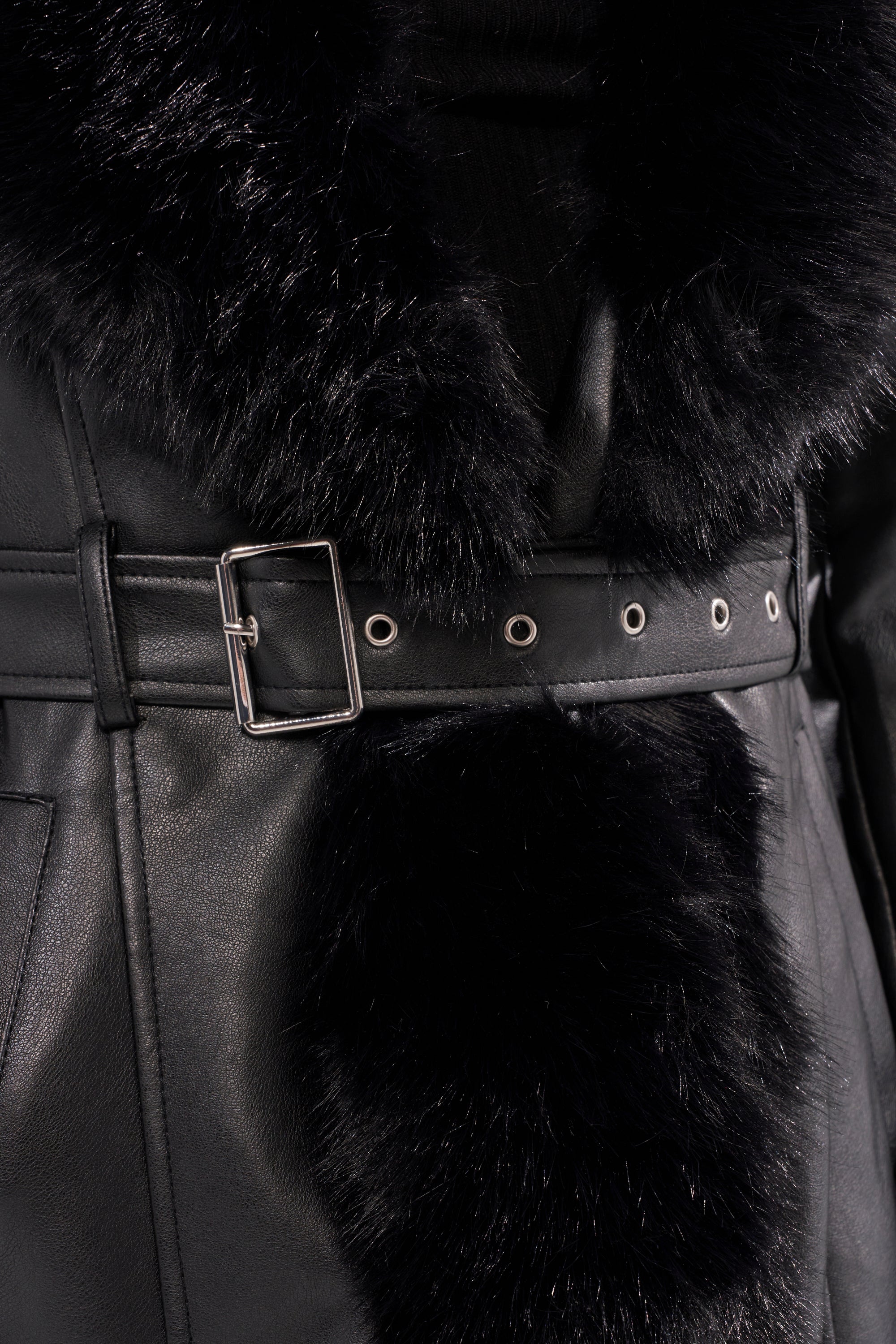 A close-up of the VIVAN LONG PU TRENCH WITH FAUX FUR TRIM, a black faux leather trench coat with a silver buckle belt and black faux fur trim along the collar and front.