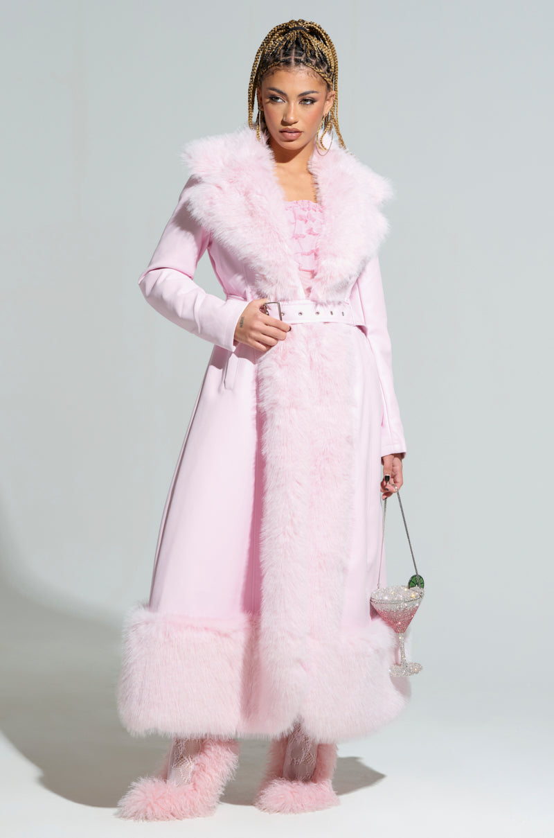 Dressed in the VIVIAN FUR TRIM COAT, an individual exudes confidence in a long pink coat accentuated by matching fluffy boots. The quilted satin lining of the coat enhances its elegance. They hold a large, ornate glass containing a pink beverage with green garnish against a plain gray background.
