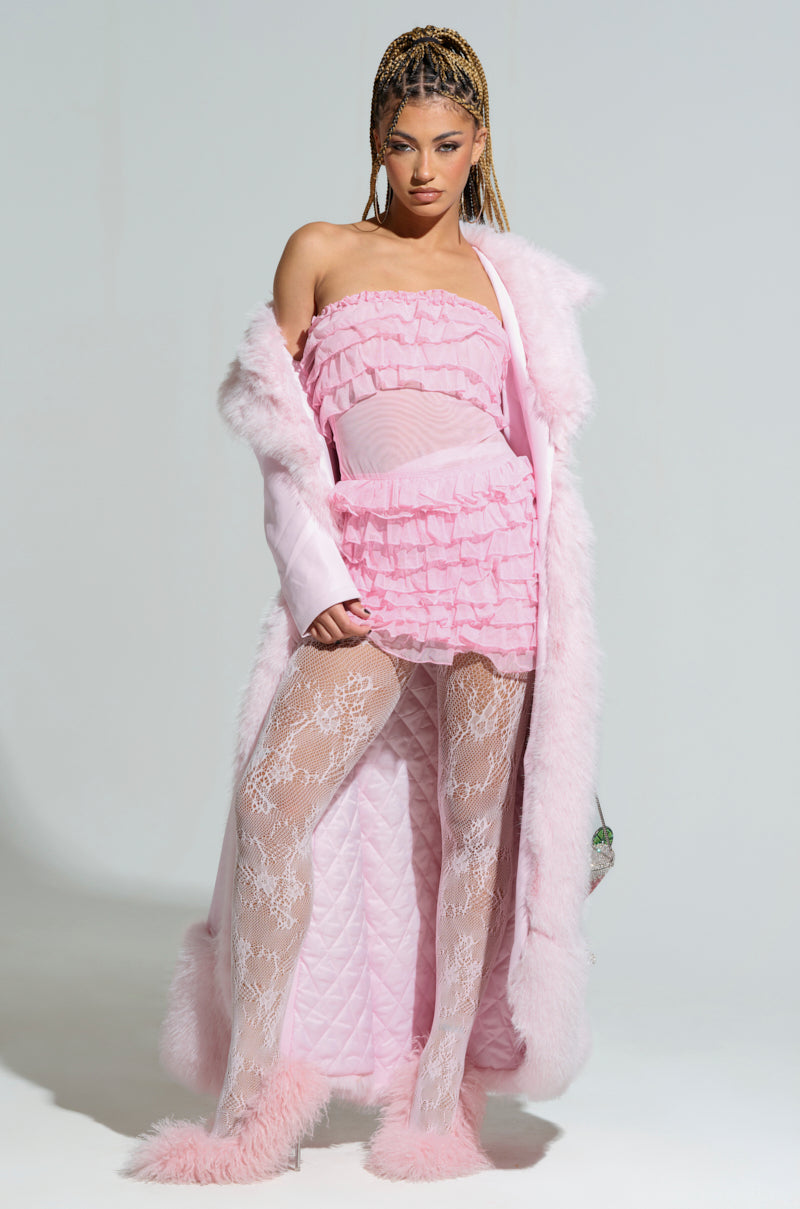 A person exudes confidence in a pink ruffled dress complemented by the VIVIAN FUR TRIM COAT and lace tights. Fluffy pink shoes complete the ensemble, while their braided hairstyle enhances their bold fashion against a plain white background.