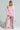 A person exudes confidence in a pink ruffled dress complemented by the VIVIAN FUR TRIM COAT and lace tights. Fluffy pink shoes complete the ensemble, while their braided hairstyle enhances their bold fashion against a plain white background.