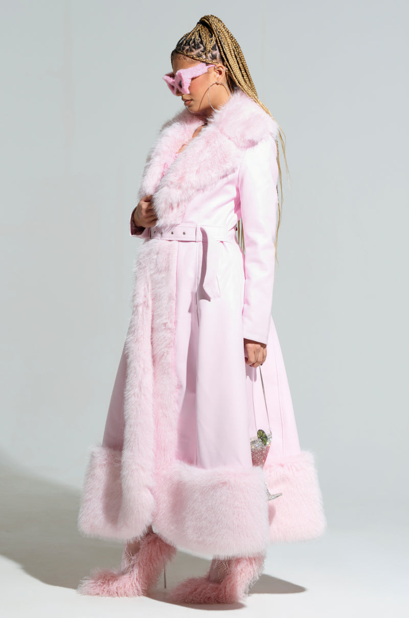 A person confidently stands wearing the VIVIAN FUR TRIM COAT, a long, light pink piece with faux fur trim and quilted satin lining. They have braided hair and are accessorized with pink sunglasses, matching furry boots, and a small purse. The plain background accentuates their stylish ensemble.