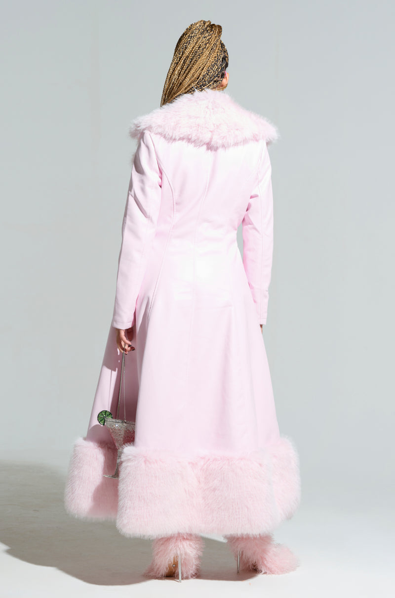 Wrapped in the VIVIAN FUR TRIM COAT, a person is adorned in a long, light pink coat featuring fluffy accents on the collar, hem, and sleeves. They accessorize their look by carrying a small bag and wearing matching fluffy ankle boots against a plain light gray backdrop.
