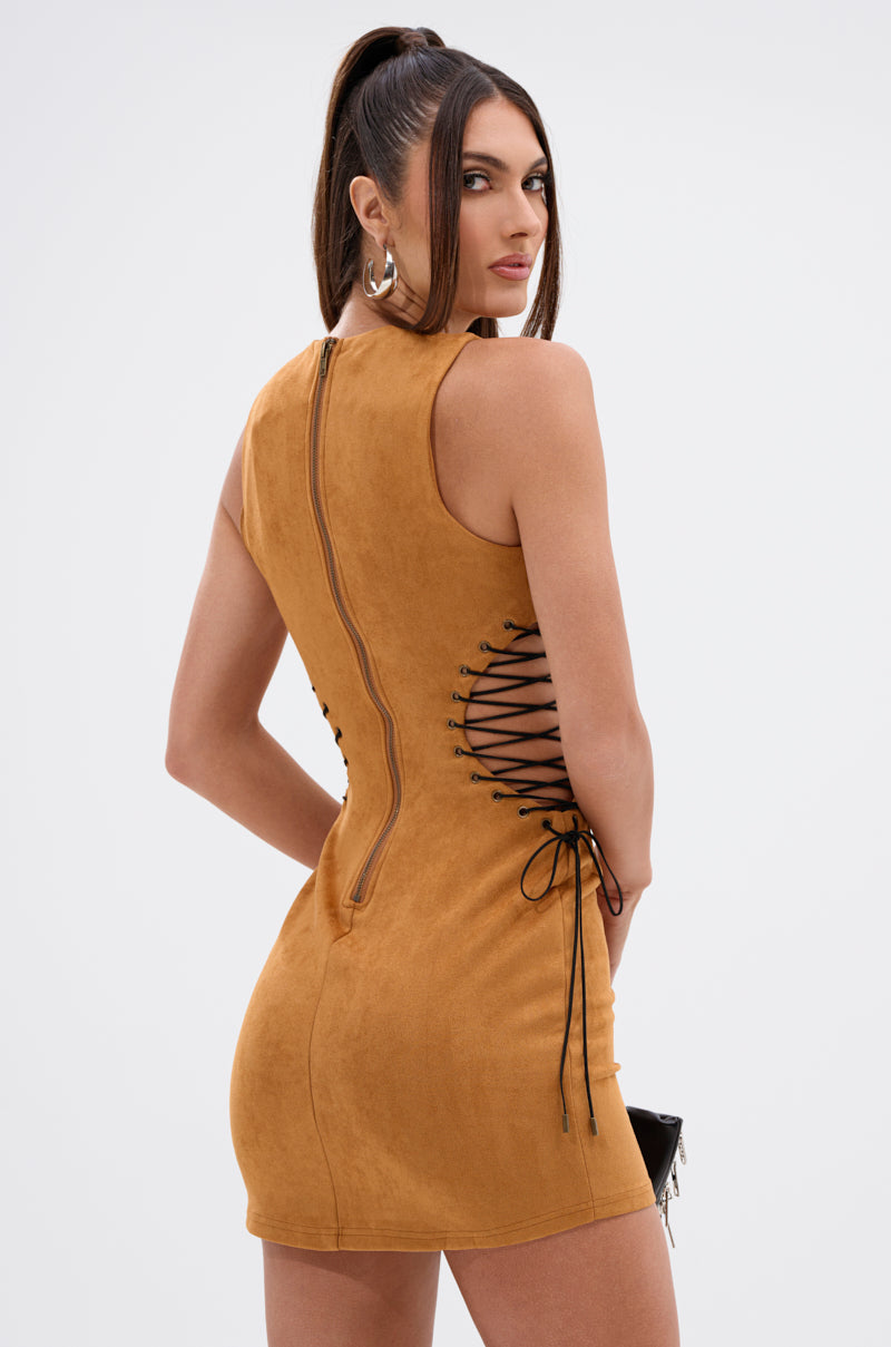 Model in THE DESERT IS CALLING SUEDE MINI DRESS, a sleeveless, curve-hugging faux suede mini with black lace-up sides. She poses with her back mostly to the camera, glancing over her shoulder and wearing her long dark hair in a high ponytail.