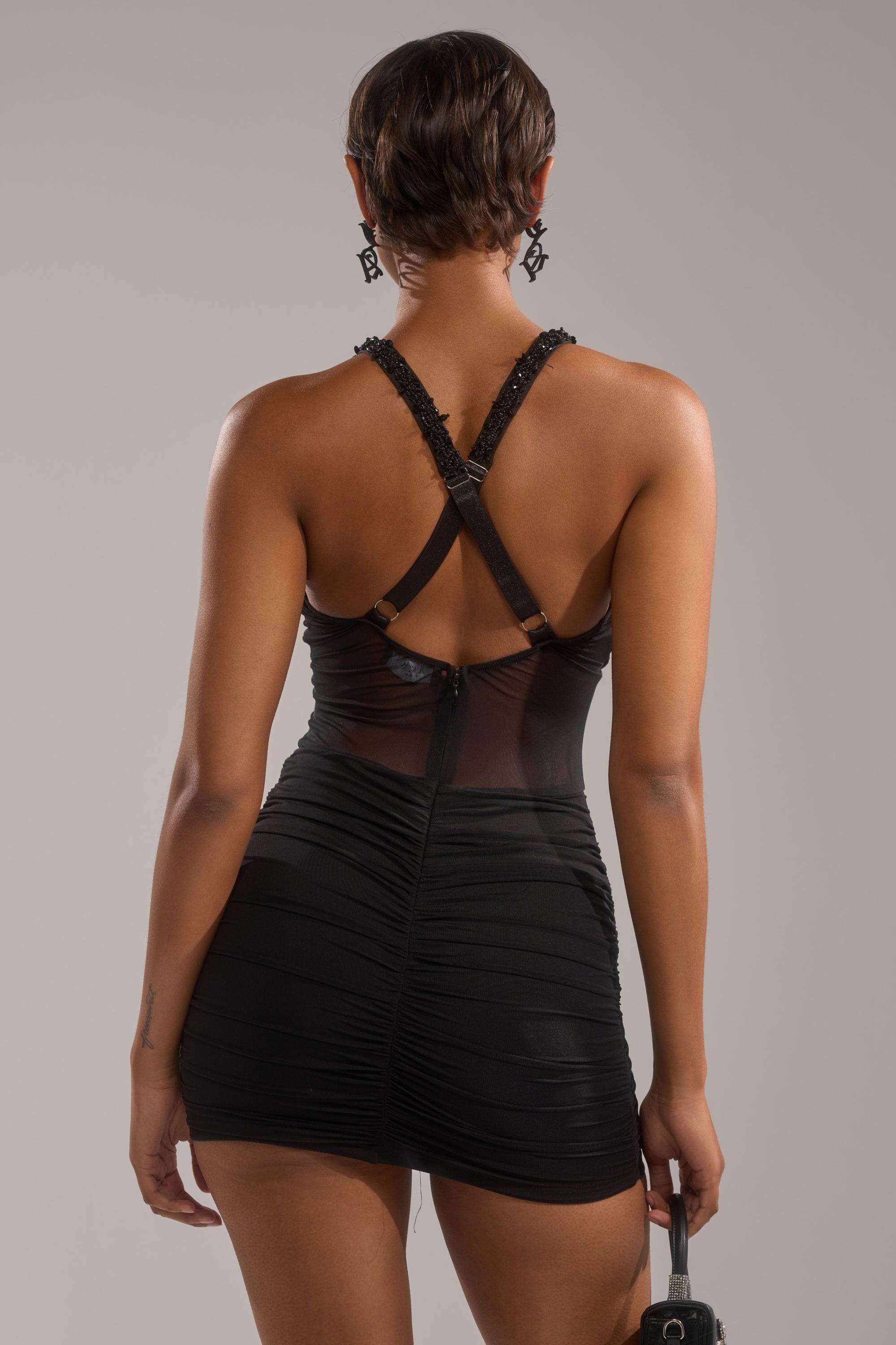 A person with short hair is seen from behind, wearing the fitted black SHAKE IT GIRL MINI DRESS by AKIRA Label with crisscross straps. They hold a small black purse and wear dangling earrings against a plain, neutral background.