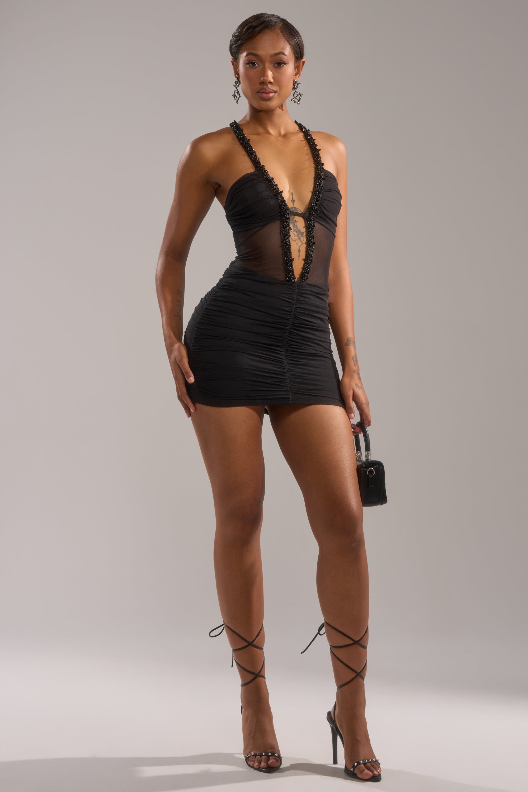 A woman poses confidently in the SHAKE IT GIRL MINI DRESS, featuring lace details and sheer panels. She completes the look with strappy black heels, a small handbag, and drop earrings against a plain gray background.