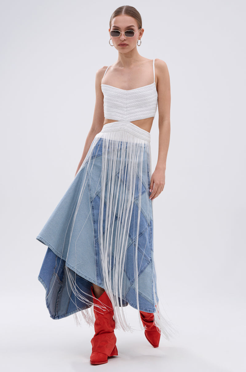 A woman showcases festival style in the RODEO READY FRINGE MAXI DRESS IN WHITE, paired with round sunglasses, hoop earrings, and red heeled boots, standing against a plain light background.