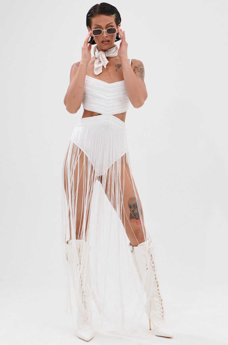 A woman models the RODEO READY FRINGE MAXI DRESS IN WHITE with a fringed skirt, white lace-up boots, sunglasses, and a neck scarf. Her arm and thigh tattoos add edge to her standout festival look against a plain white background.