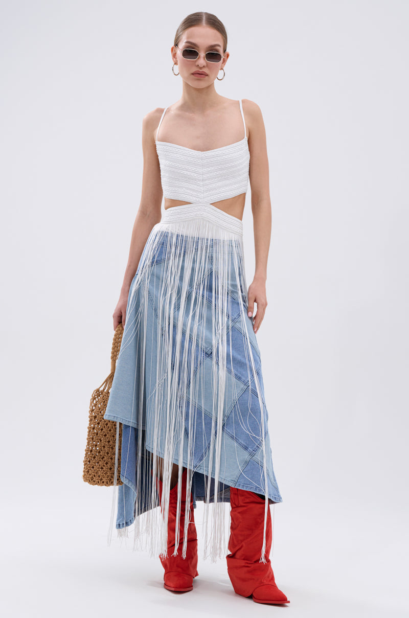 A woman stands against a plain backdrop wearing sunglasses, hoop earrings, red boots, a woven brown bag, and the Rodeo Ready Fringe Maxi Dress in White—her look is the epitome of Western-inspired festival style.