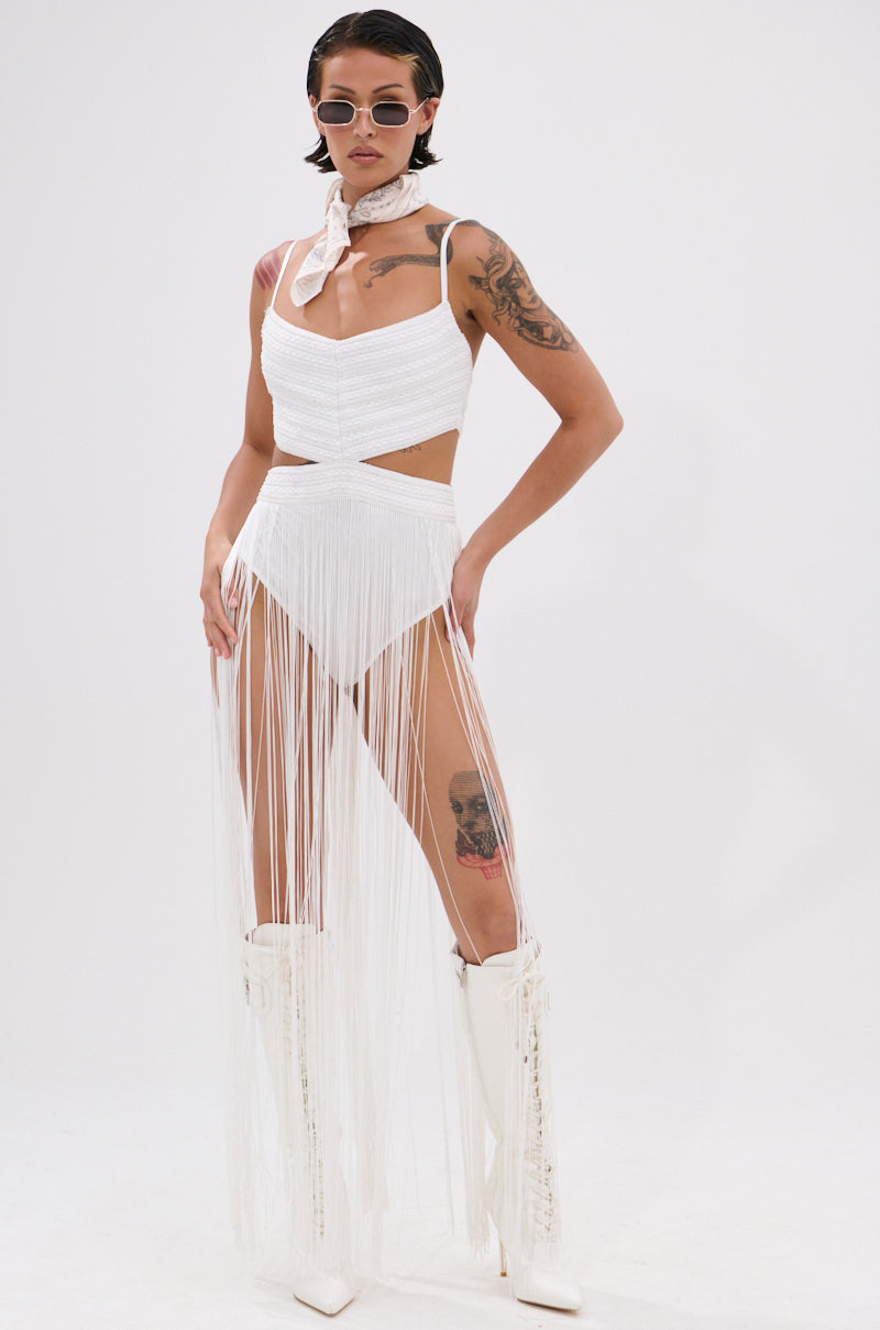 A person in the Rodeo Ready Fringe Maxi Dress in White, paired with white knee-high boots, a neck scarf, and round sunglasses stands confidently against a white background—serving bold festival style with short dark hair and a thigh tattoo.