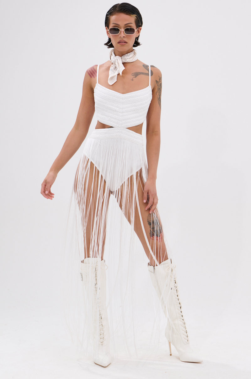 A woman models the RODEO READY FRINGE MAXI DRESS IN WHITE with high boots, a bandana, sunglasses, and visible tattoos—embracing festival style against a plain white background.