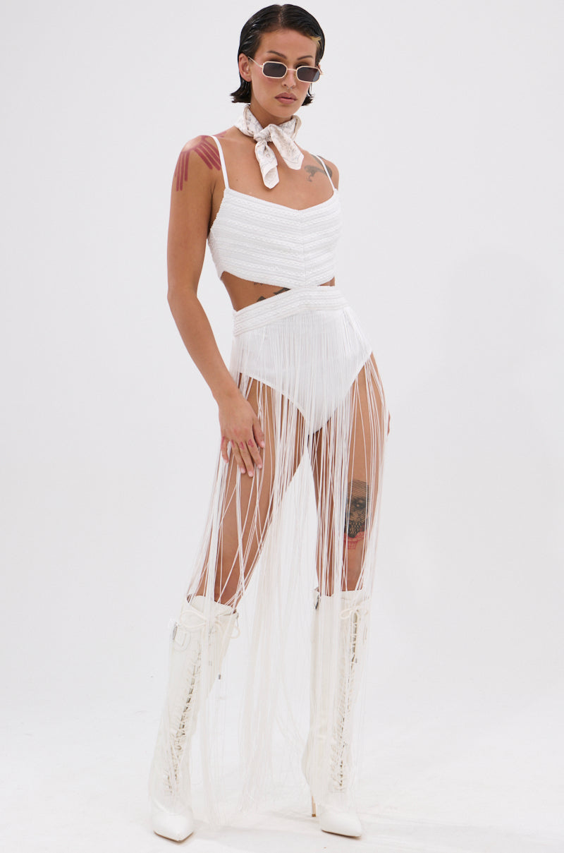 A woman embraces festival style in the RODEO READY FRINGE MAXI DRESS IN WHITE, paired with a textured crop top, high-waisted bottoms, knee-high boots, sunglasses, a neck scarf, and showing her arm and thigh tattoos.