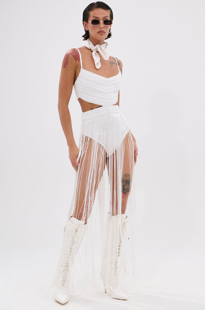 Confidently standing against a plain white background, a person wears sunglasses, a white scarf, knee-high white lace-up boots, and the RODEO READY FRINGE MAXI DRESS IN WHITE—an ideal look for festival fashion.