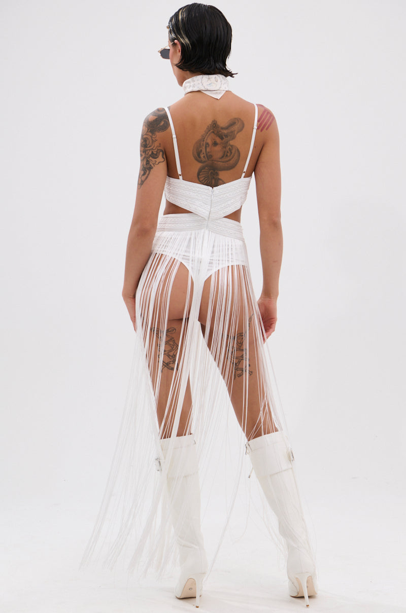 Model faces away in the RODEO READY FRINGE MAXI DRESS IN WHITE, styled with knee-high boots, sunglasses, and a bandana. Tattoos on arms, back, and legs plus short dark hair complete this bold festival look against a plain white background.