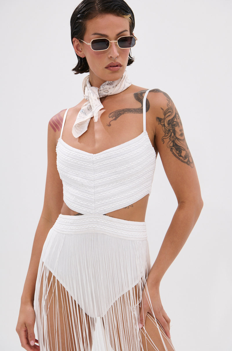 A person in the Rodeo Ready Fringe Maxi Dress in White, sunglasses, and a patterned neck scarf stands against a plain background, showing off short, slicked-back hair and visible arm and shoulder tattoos.