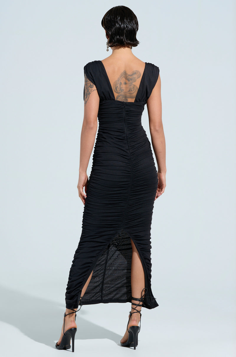 A person with short dark hair stands with their back to the camera, showcasing a TIME TO PARTY MIDI DRESS in black. The dress is ruched and bodycon, featuring a slit at the hem. They pair it with strappy high heels and display a large tattoo on their upper back.