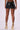 A person stands facing away, showcasing the BUCKLE UP MINI SKIRT, a fitted faux leather mini skirt with an embossed snakeskin pattern. They are wearing high heels with gold straps that wrap around their ankles and lower legs. The background is a plain, light-colored surface.