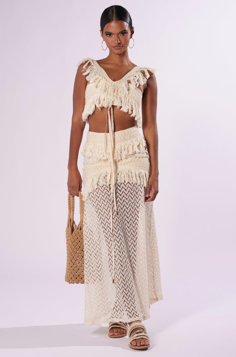 A woman showcases a boho-chic ensemble, wearing the ON THE FRINGE SKIRT, which is high-waisted and sheer with a zigzag pattern. She pairs it with a light-colored, fringed crop top and completes her look with a woven handbag and sandals. Her pulled-back hair is accentuated by hoop earrings, while the plain white background highlights her festival-ready outfit.