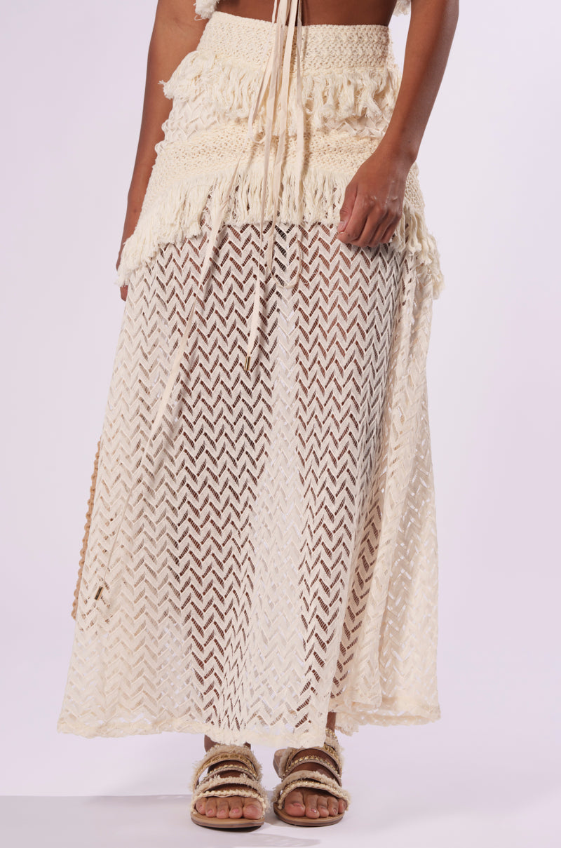 A person wearing the ON THE FRINGE SKIRT, a bohemian-style beige maxi skirt featuring a sheer, intricate zigzag pattern overlay and textured waist detail, perfect for your festival wardrobe. Complementing the outfit are beige sandals. The background is plain and neutral.